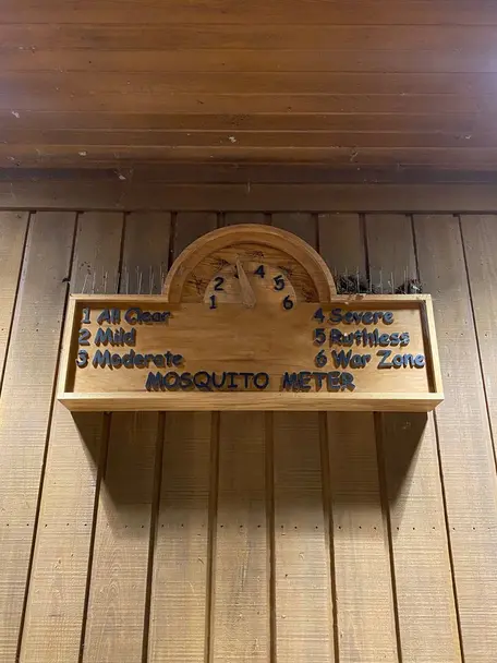 Wooden sign called the Mosquito Meter, with a scale from 1 - 6, with the scale hand resting on 3, where 1 represents All Clear, 2 = Mild, 3 = Moderate, 4 = Severe, 5 = Ruthless, and 6 = War Zone.