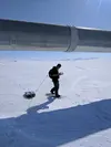 A person wearing snowshoes pulls a ground penetrating radar unit across an ice field using a rope tethered to their backpack. They are walking near a large pipeline and are holding a monitor to view incoming data.