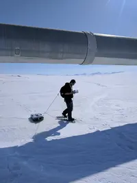 A person wearing snowshoes pulls a ground penetrating radar unit across an ice field using a rope tethered to their backpack. They are walking near a large pipeline and are holding a monitor to view incoming data.