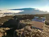 A small box-shaped gravimeter sits on rocks in the foreground while a volcano shrouded in clouds is visible in the background.