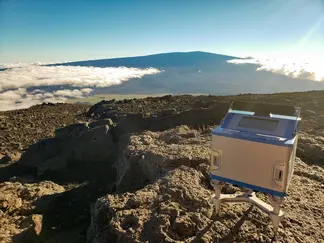 A small box-shaped gravimeter sits on rocks in the foreground while a volcano shrouded in clouds is visible in the background.