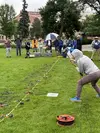 A seismic refraction survey is conducted in a grassy field of a college campus. A tape measure is stretched across the ground and geophones are placed at even intervals. In the foreground, someone swings a sledgehammer towards a metal plate on the ground. In the background, a group of people at the end of the tape measure monitor data collection with a laptop and other equipment.