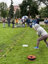 A seismic refraction survey is conducted in a grassy field of a college campus. A tape measure is stretched across the ground and geophones are placed at even intervals. In the foreground, someone swings a sledgehammer towards a metal plate on the ground. In the background, a group of people at the end of the tape measure monitor data collection with a laptop and other equipment.