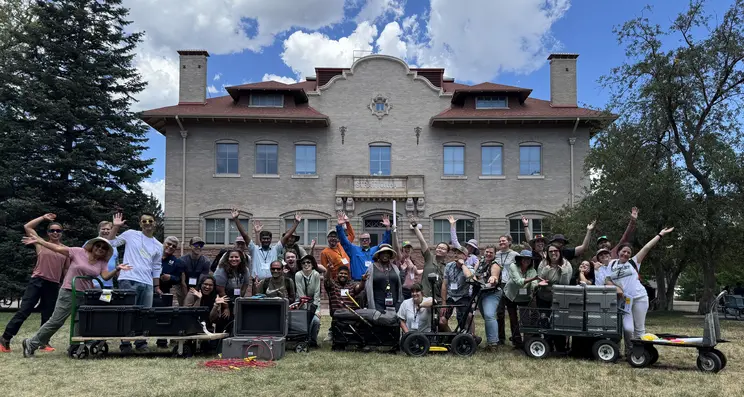 Group of ~32 people in  front of one of the older Colorado School of MINES buildings along with some geophysics instruments on carts.