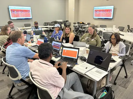 A team of 10 people around a large table in a classroom working on laptops. GPR data showing on various laptop and classroom screens.