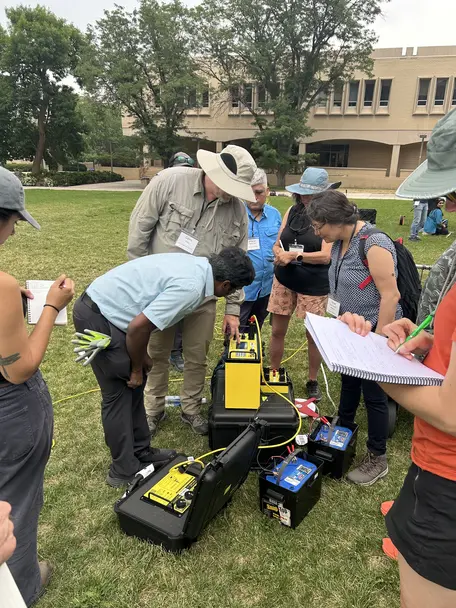 Helf a dozen adults grouped around an electrical resistivity instrument on a campus quad.