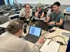 People grouped around two laptops at a table in a classroom.