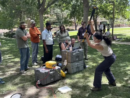 A woman swings a sledge hammer in the foreground to send a seismic wave. Another half dozen workshop particapant and grouped around a laptop set on stacked packing crates running the survey software and watching.