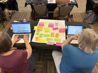 Two project authors working on their laptops which refering to a large post-it sheet of brainstorming ideas.