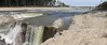 Waterfalls in foreground to the left along a newly cut river channel, with remains of a breached earthen dam to the back right. 