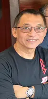 Dr. Joel Aquino looks slightly upward beyond the camera focus, smiling and listening intently. He is wearing a black T-shirt with a partially visible red logo, has short dark hair, wears glasses, and has his arms casually crossed