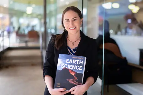A woman smiling and holding a textbook that says Earth Science in large letters on the cover